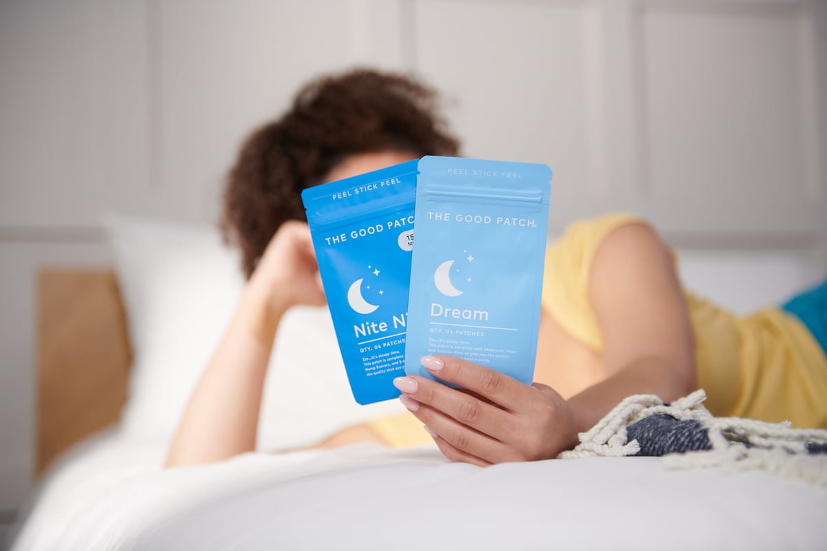Model covering her face by holding two The Good Patch products while on bed