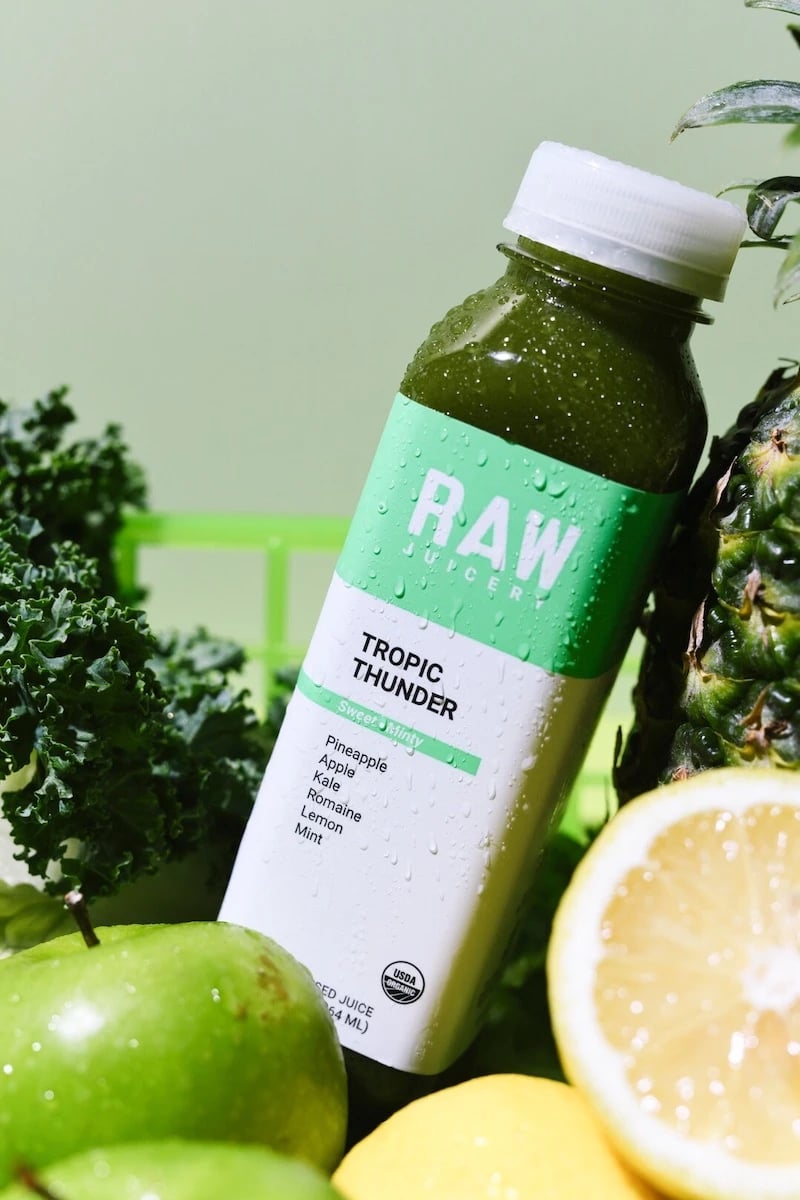 Raw Juicery bottle leaning on pineapple and surrounded by fruits and vegetables